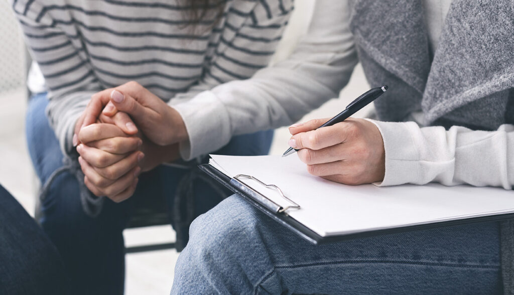 A therapist comforts a client while taking progress notes on a clipboard.